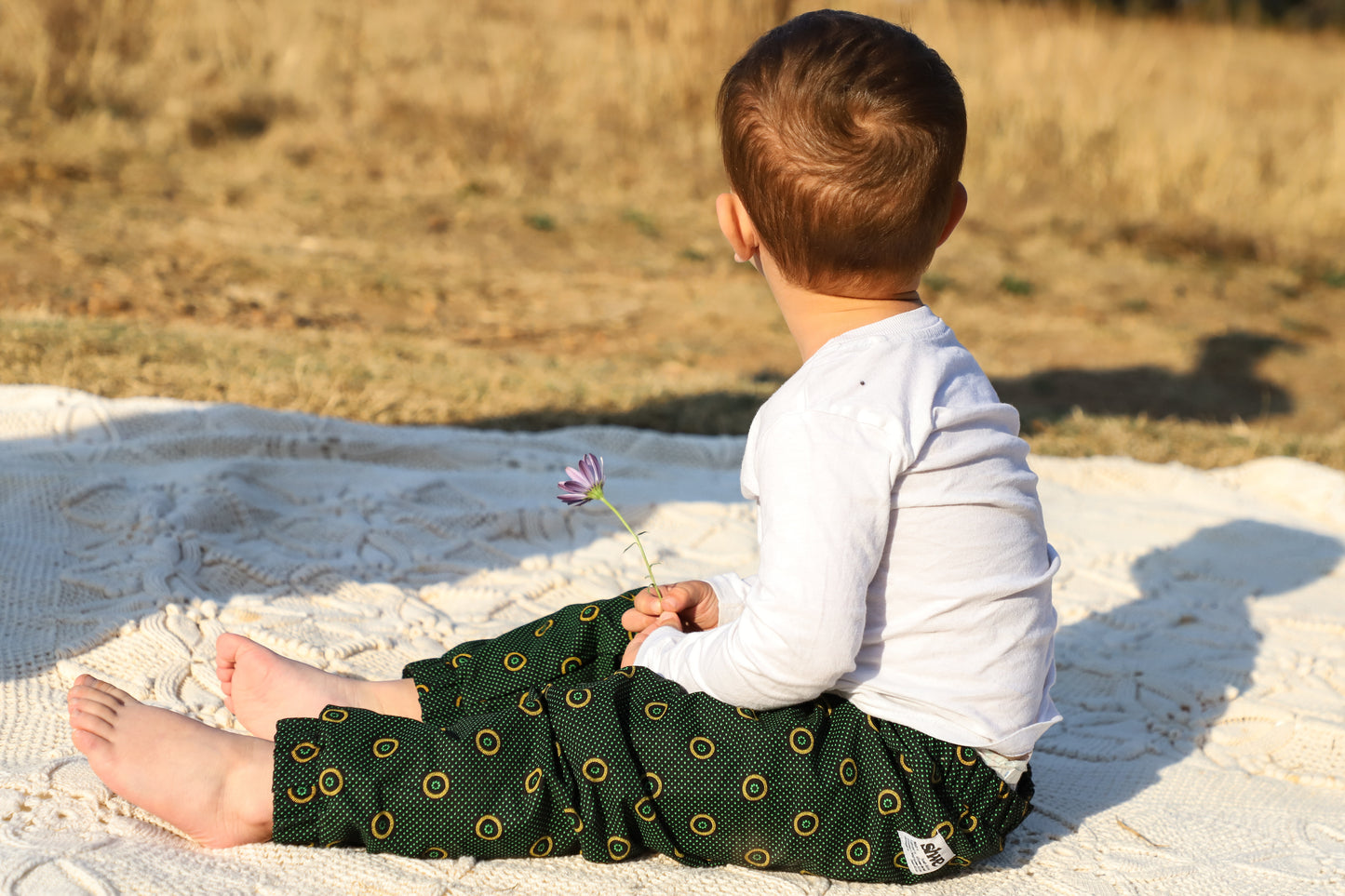 un petit garçon assis sur une couverture avec un pantalon imprimé shweshwe vert avec une fleur dans les mains