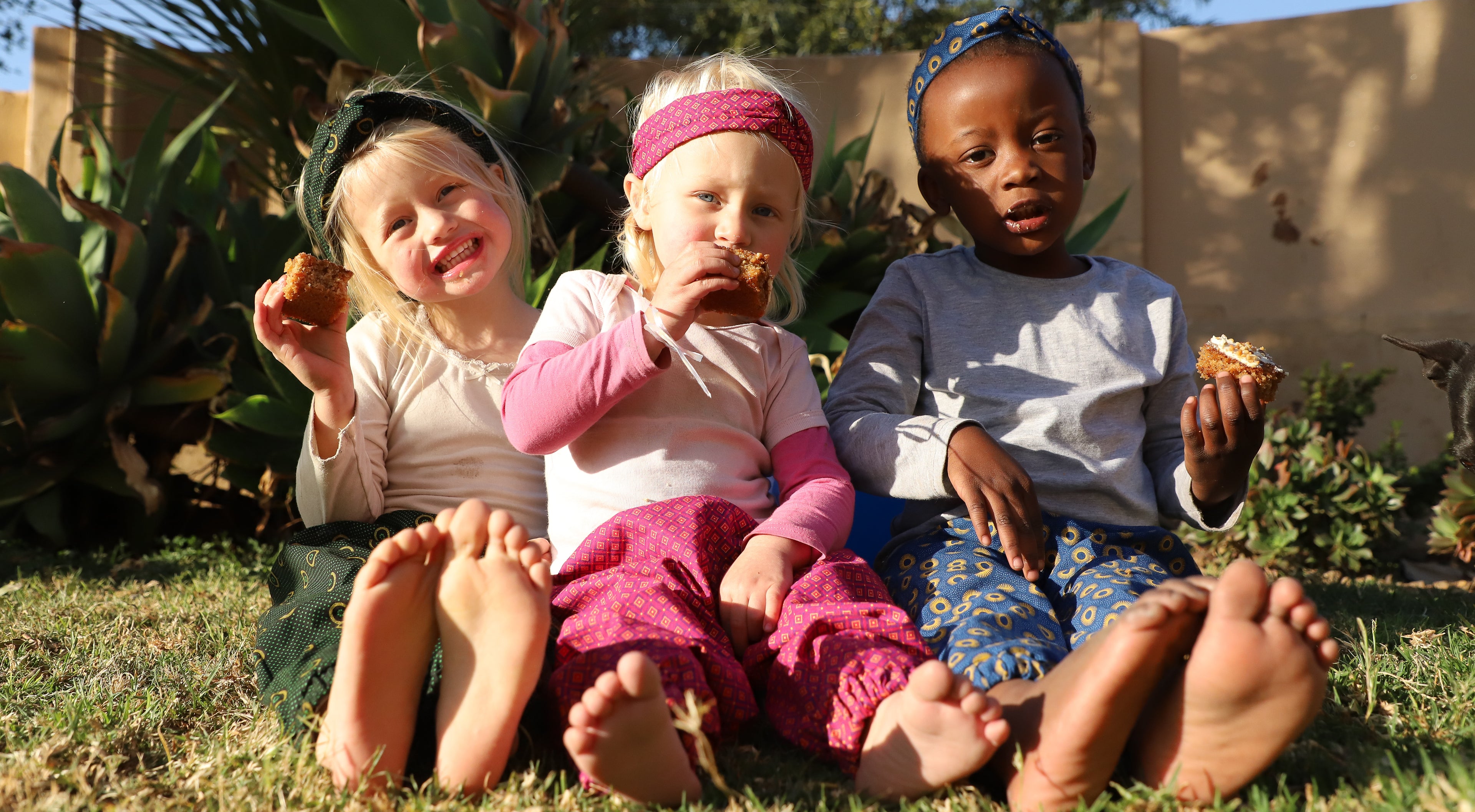 From left to right: white toddler in green african print trousers and headband, white toddler in pink african print trousers and headband, black toddler in blue african print trousers and headband.