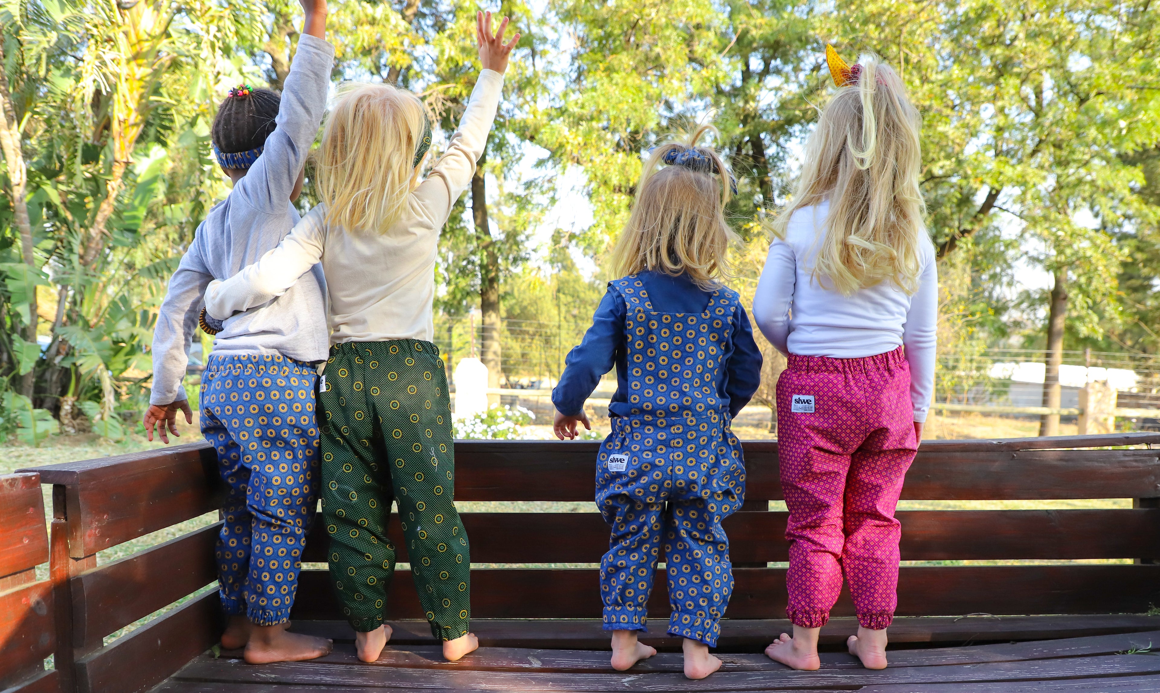 From left to right: black child in blue african print pants, white child in green african print pants, white child in blue african print dungarees, white child in pink african print pants. African print is swheswhe fabric. Ethically made in South Africa. 